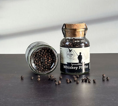 Whiskey Pfeffer im Korkglas. Vor dem Glas liegen Pfefferkörner. Links danebn liegt ein Glas Wiskey Pfeffer, zu sehen ist die Unterseite mit dem Inhalt bestehend aus Pfefferkörnern.