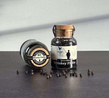 Whiskey Pfeffer im Korkglas. Vor dem Glas liegen Pfefferkörner. Links danebn liegt ein Glas. Zu sehen ist der Korkversachluss mit der Banderole.